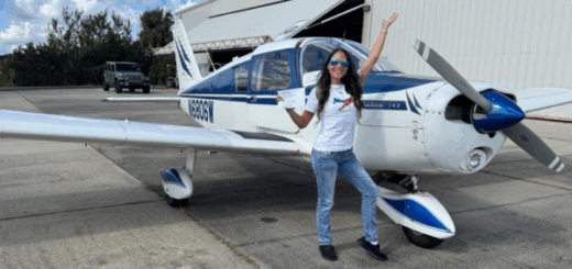 A woman holding her private pilot certificate standing in blue jeans, a white t-shirt, and aviator style sunglasses stands in front of a Piper Cherokee with her hand in the air. Rebecca Williams of the Florida Firstcoast
