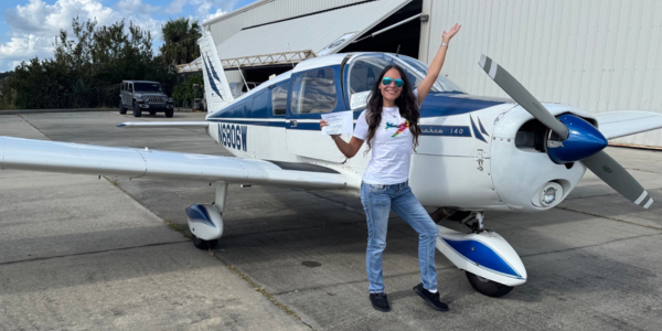 A woman holding her private pilot certificate standing in blue jeans, a white t-shirt, and aviator style sunglasses stands in front of a Piper Cherokee with her hand in the air. Rebecca Williams of the Florida Firstcoast