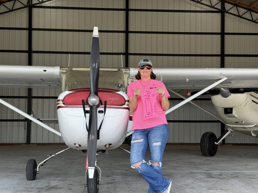 A woman in a pink shirt is holding up the back of her shirt, which has been cut off and drawn on with the airport runway drawn on, and information about her first flight. She is standing in front of a Cessna high wing training airplane.