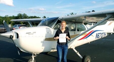 A woman stands in front of a high wing airplane holding her pilot's certification.
