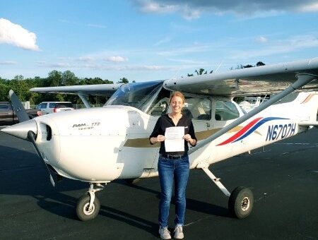 A woman stands in front of a high wing airplane holding her pilot's certification.