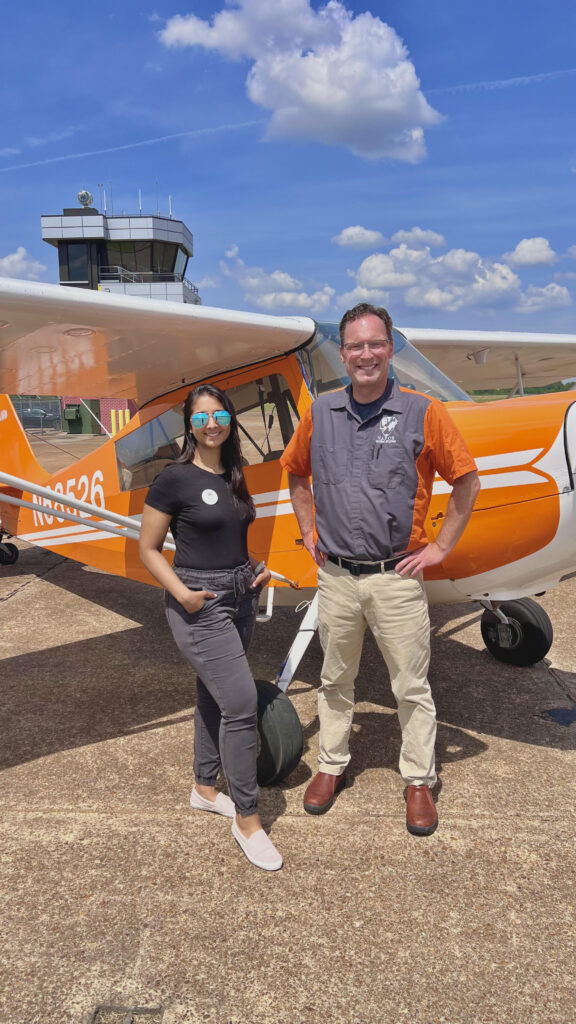 A woman and man stand in front of an orange and white 1972 Bellanca Citabria 7ECA on a sunny day with an airport control tower in the background.