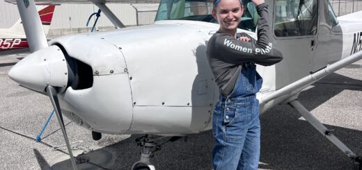 Young female pilot stands in front of a highwing airplane with her arm raised to show her strength and success.