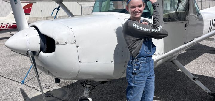 Young female pilot stands in front of a highwing airplane with her arm raised to show her strength and success.
