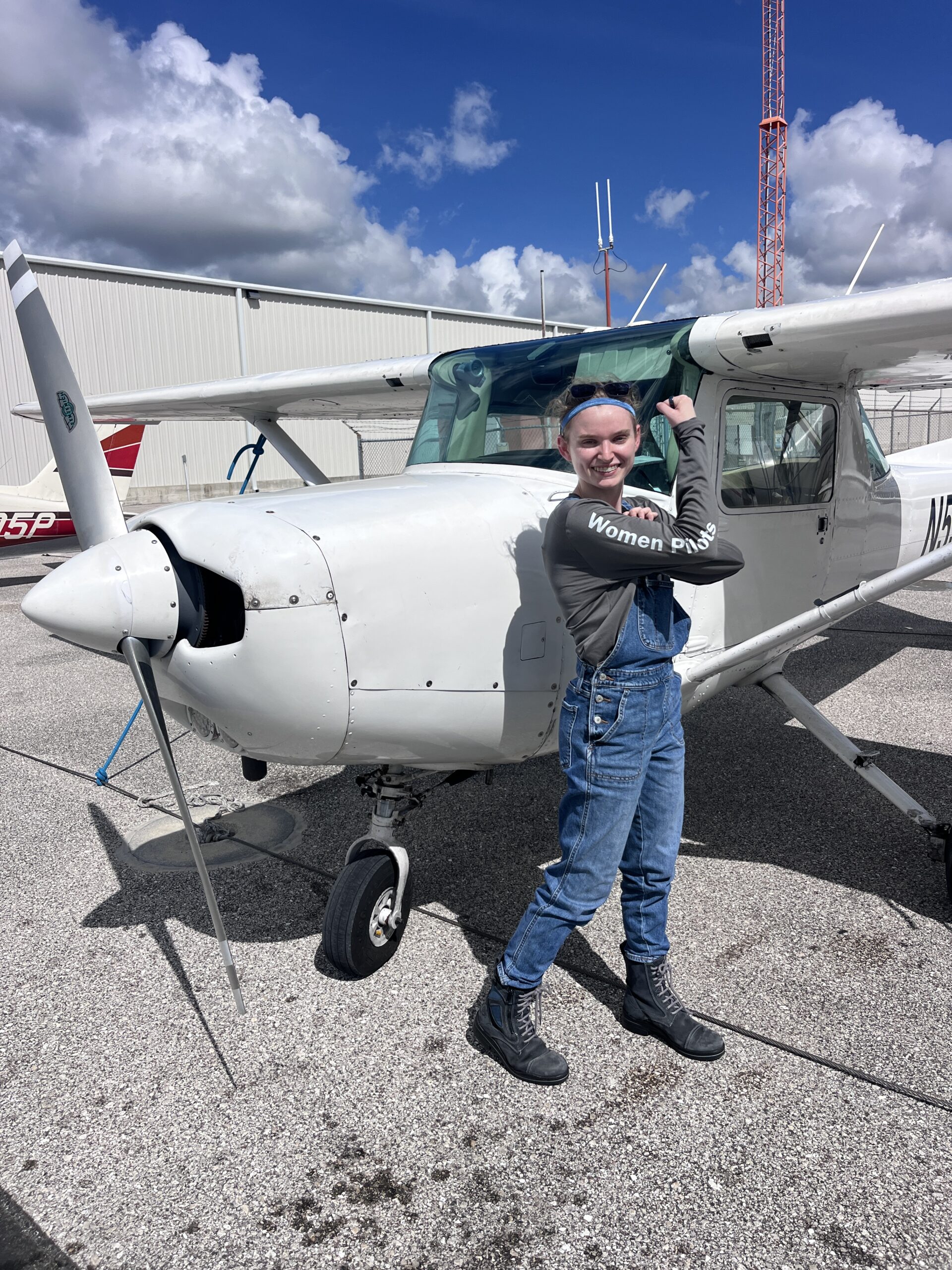 Young female pilot stands in front of a highwing airplane with her arm raised to show her strength and success.
