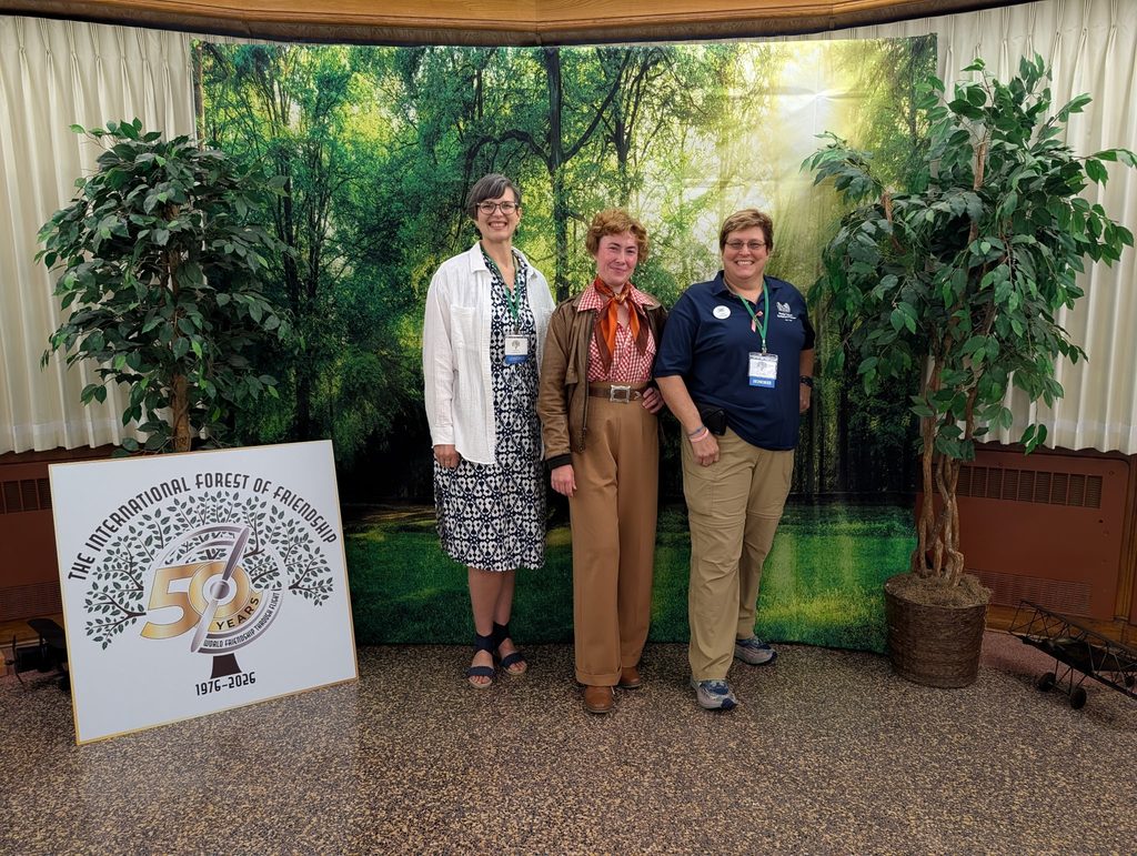 Mandi Hill, an Amelia Earhart reenactor, and Lois Horne stand together in front of a backdrop of the Forest of Friendship and adjacent to an event sign.