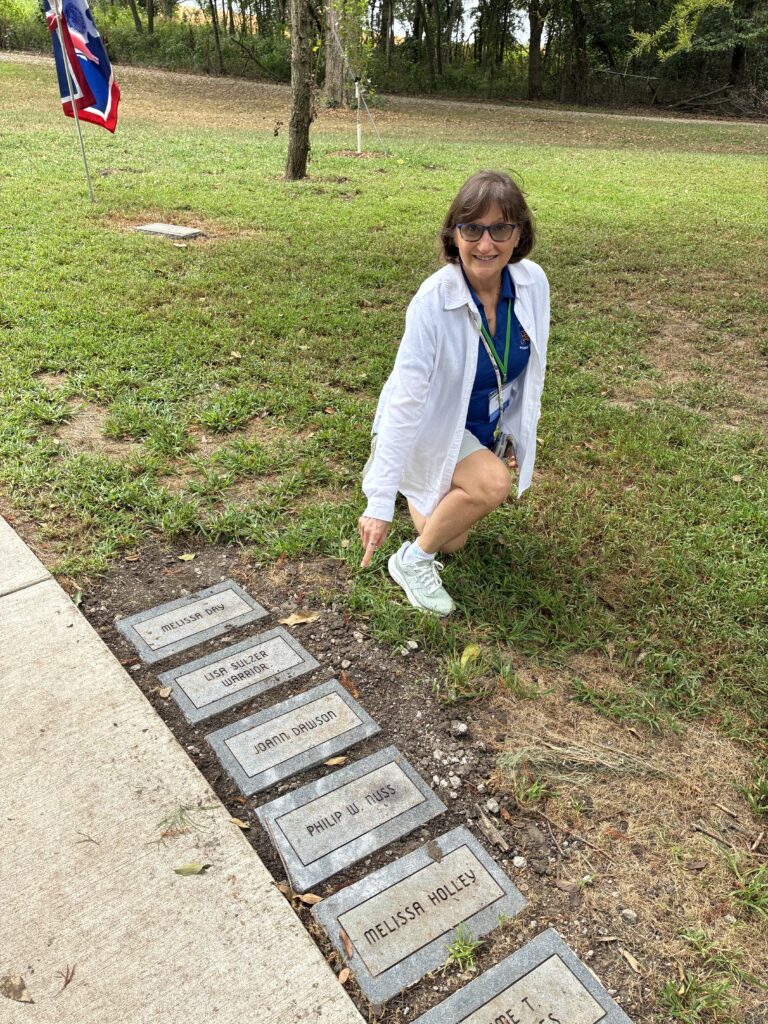 JoAnn Dawson kneels by and points to the plaque with her name along the Forest of Friendship walkway.